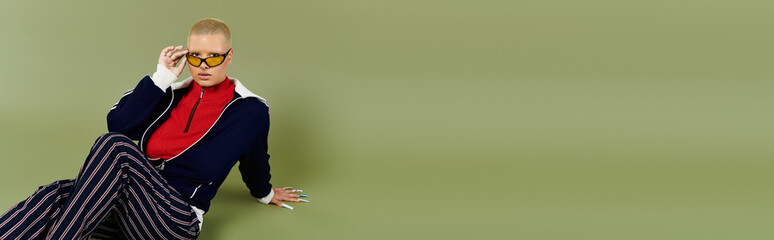 In a bold outfit, a young woman adjusts her sunglasses while sitting confidently on the ground.