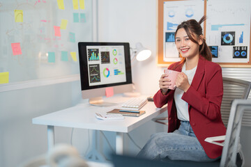 Young businesswoman is holding a mug and smiling while sitting at her desk in a modern office.  She...