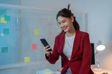 Young businesswoman is checking her smartphone and smiling while working late in a modern office. ...
