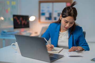 Determined businesswoman works late in her office, using a calculator and laptop for finance tasks. She's focused, analyzing data and planning for the future in a serene blue-lit atmosphere