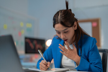 Asian businesswoman looks shocked while taking notes at a corporate meeting in a modern office, analyzing financial data on a laptop