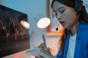 Focused stockbroker works late into the night, analyzing market data and talking on her headset in a modern office setting