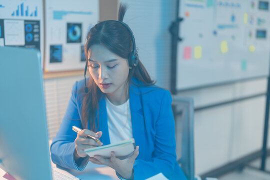 Asian businesswoman works late in her office, multitasking during a conference call, focused on her computer screen in a modern corporate setting