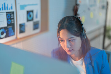 Focused businesswoman works late at night in the office, using a computer and headset for a conference call, showing dedication. Blue lighting adds calmness to the busy atmosphere