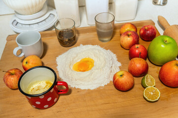 Wooden kitchen board with many ingredients (flour,egg,apple,roll,nelted butter in red mug,glass,raisin,jar) for preparation of baking apple strudel.