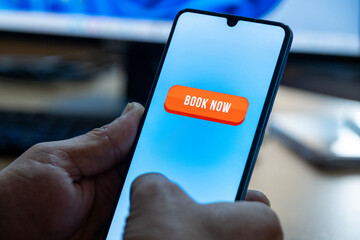Close-up of the “Book Now” message on the smartphone screen. Online booking concept. Office background. Early booking message. 