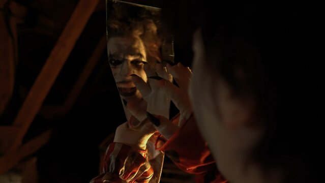 Crazy creepy killer man dressed as clown with makeup at attic in ruined house holding mirror shard looks in it crazy smiling. Halloween thriller concept.