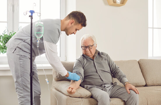 Young friendly nurse help gray-haired sick senior elderly man in receiving IV drip infusion and vitamin injection therapy sitting on sofa at home or in medical clinic. Health care concept.