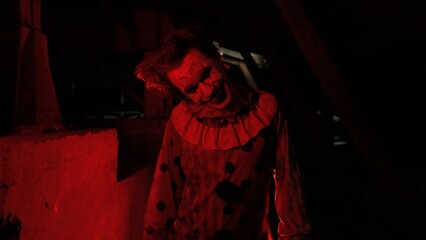 Crazy creepy killer man dressed as clown with makeup at attic in ruined house looks at camera, talking, smiling going crazy. Halloween thriller concept.