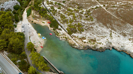 Aerial view of a cave in a cove on a stretch of rocky coast. There are pedal boats and canoes on the beach.
