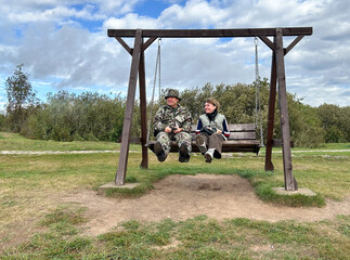 Joyful elderly couple sitting on a wooden swing in their backyard, surrounded by a wooden fence and...