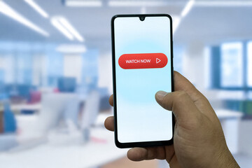 “Watch Now” message on close-up smartphone screen. Watching online video. Office background. Video streaming platform, video advertising and streaming concept.