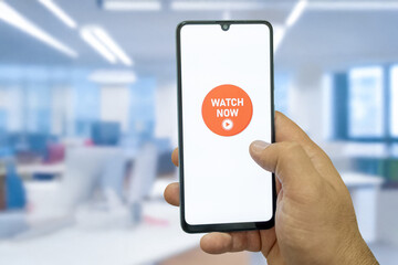 “Watch Now” message on close-up smartphone screen. Watching online video. Office background. Video streaming platform, video advertising and streaming concept.