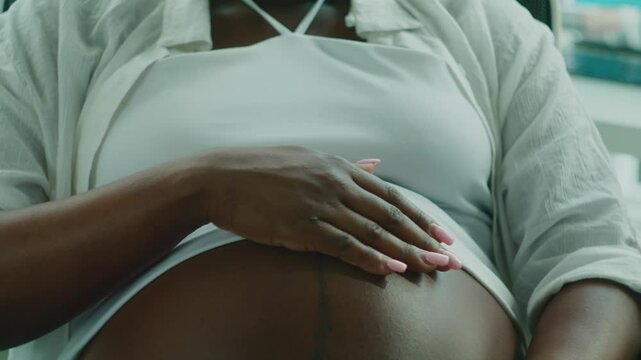 Close-up of an Afro-Latin pregnant woman gently rubbing her belly, showing a tender and intimate moment. Ideal for themes of pregnancy, self-care, and maternal love in a cozy home setting.