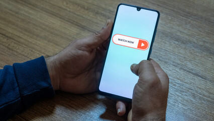 “Watch Now” message on close-up smartphone screen. Watching online video. Office background. Video streaming platform, video advertising and streaming concept.