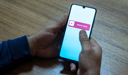 “Watch Now” message on close-up smartphone screen. Watching online video. Office background. Video streaming platform, video advertising and streaming concept.