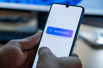 “Watch Now” message on close-up smartphone screen. Watching online video. Office background. Video streaming platform, video advertising and streaming concept.