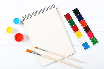 Colorful gouache paints with brushes and blank sheet of paper on white wooden table