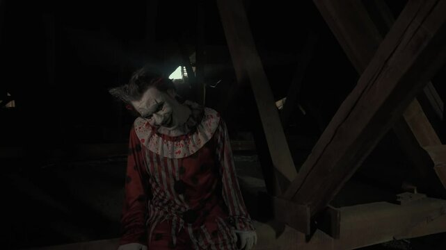 Crazy creepy killer man dressed as clown with makeup at attic in ruined house sits on wooden beam swinging legs and smiles. Halloween thriller concept.