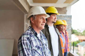 Senior contractor standing by coworkers wearing hardhats looking out from incomplete house