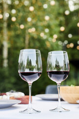 Vertical closeup image of two red wine glasses on festive dinner table outdoors set for party occasion with greenery in background