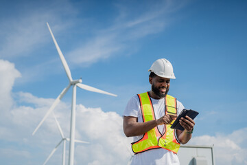 A man in a yellow vest is looking at a tablet while standing next to a wind turbine. electrical, engineer, environment, energy, clean, sustainable, wind turbine, nature