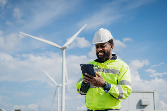 A man in a yellow safety jacket is looking at a tablet while standing near a wind turbine. electrical, engineer, environment, energy, clean, sustainable, wind turbine, nature