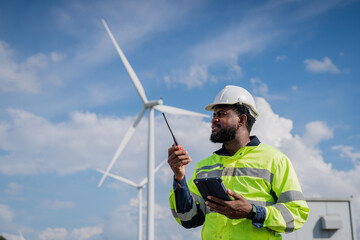 A man in a yellow safety vest is holding a walkie talkie and looking up at a wind turbine....