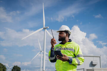 A man in a yellow safety vest is standing next to a wind turbine and holding a walkie talkie. electrical, engineer, environment, energy, clean, sustainable, wind turbine, nature