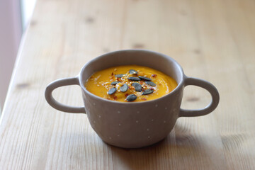 Bowl of creamy pumpkin and potato soup, with pumpkin seeds and chilli flakes toppings. Selective focus, wooden background.