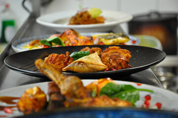 Crispy samosas and succulent chicken pieces, garnished with fresh greens and a sprinkle of red spices. In the background, blurred plates of colorful exotic foods create a bustling atmosphere.