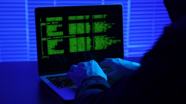 Hooded hacker using laptop computer, close-up. Anonymous hacker sitting in the dark office, typing command on computer monitor