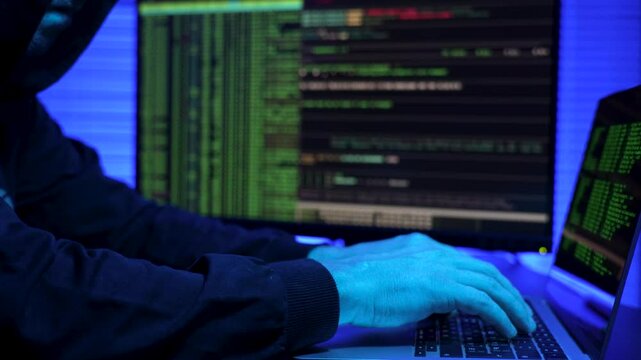 Hooded hacker using laptop computer, close-up. Anonymous hacker sitting in the dark office, typing command on computer monitor