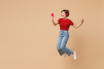 Full body young happy cool woman she wear red t-shirt casual clothes jump high hold in hand use mobile cell phone listen to music in headphones isolated on plain beige background. Lifestyle concept.
