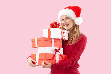Cheerful woman holding many red Christmas present gift boxes while wearing Santa hat, isolated on pink background. Perfect for sale offer holiday promotions, online shopping adv banner, or gift card