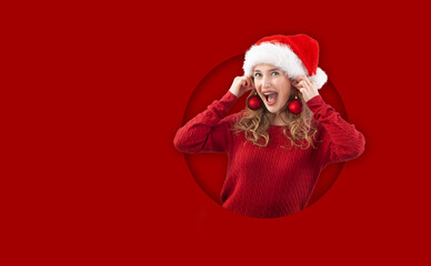 cheerful blonde young woman while wearing a Santa hat, play with christmas balls pretending earrings, isolated on red background. Template For sale and offer promotions or online shopping adv banner