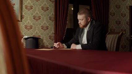 Waist up shot of concentrated 19th century gentleman wearing black suit sitting at table with red tablecloth on it in old-fashioned well-furnished room and writing letter on parchment paper