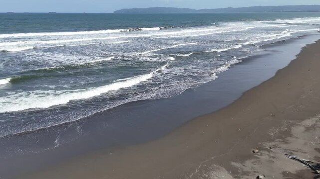 Waves and black sand at Kemiren Beach Cilacap with a background of blue sea and blue sky, 4K