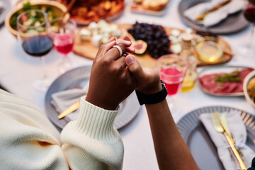 Close up of two people holding hands at dinner table and saying grace during Thanksgiving celebration with friends and family copy space