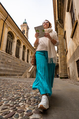 Vacation in Italy. Beautiful middle-aged woman wearing beige sweater, turquoise skirt and scarf walking with city map along cobblestone street and sightseeing old city of Bergamo on autumn day.