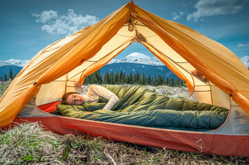 Young woman sleeping under a down blanket in a yellow tent in the morning mountains.