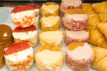 Rows of round pastries with red, yellow, and pink frosting on display next to croissants.