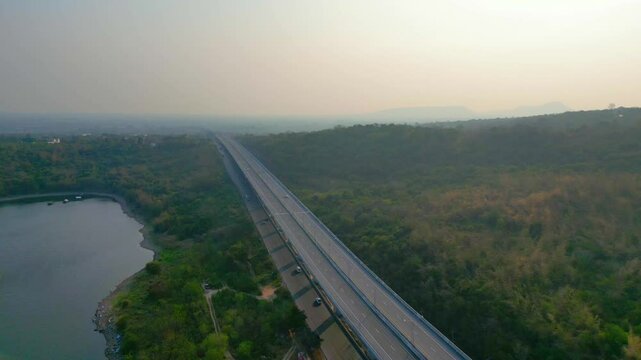 Aerial view of a highway winding through a hilly, forested landscape with wind turbines in the distance, near a coastline.

