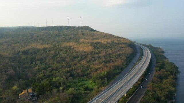 Aerial view of a highway winding through a hilly, forested landscape with wind turbines in the distance, near a coastline.

