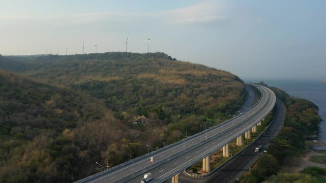 Aerial view of a highway winding through a hilly, forested landscape with wind turbines in the distance, near a coastline.
