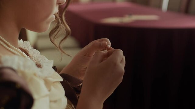 Medium closeup of beautiful young aristocratic woman in elegant 19th century attire trying to thread needle while sitting alone in well-furnished parlor room with red tablecloth and classical decor