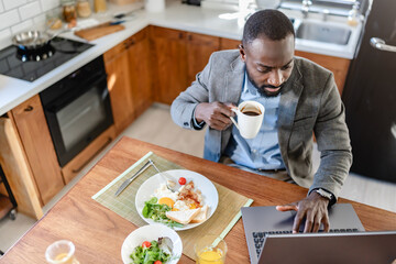 African-American man in a modern kitchen, drinking coffee and working on a laptop during breakfast. Bright and productive morning scene.