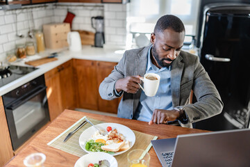 African-American man working on a laptop while eating breakfast at home. He is multitasking in a bright, modern kitchen, combining work and a healthy meal.