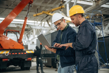 Reading information from laptop. Two male factory workers are in the warehouse
