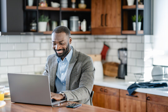 Focused African-American man working on his laptop at home, dressed in business attire. He appears engaged and concentrated in a modern kitchen workspace.